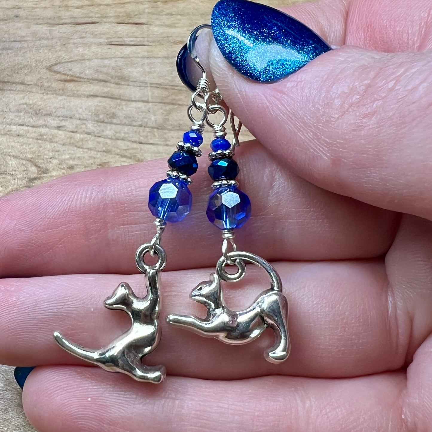 A pair of asymmetric earrings made from silvery kitty charms and three faceted glass beads in various shades of dark blue. The beads are graduated in size with thin silver spacers between them and the kitty charms swing freely below the beads. One kitty on its hind legs reaching towards the beads with its front paws and the other is doing BIG STRETCH.
In this photo they are being held in a woman’s hand. She has pale skin and sparkly blue nails.