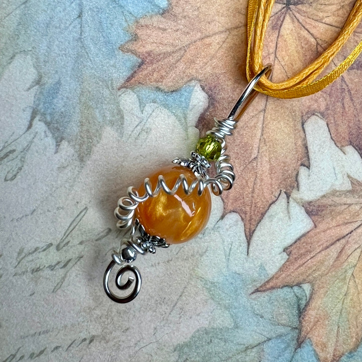 A pendant that looks like a pumpkin, made from a shimmery orange bead topped with a smaller green bead. There is a long tendril of wire twisted around it and it is on an orange ribbon. 