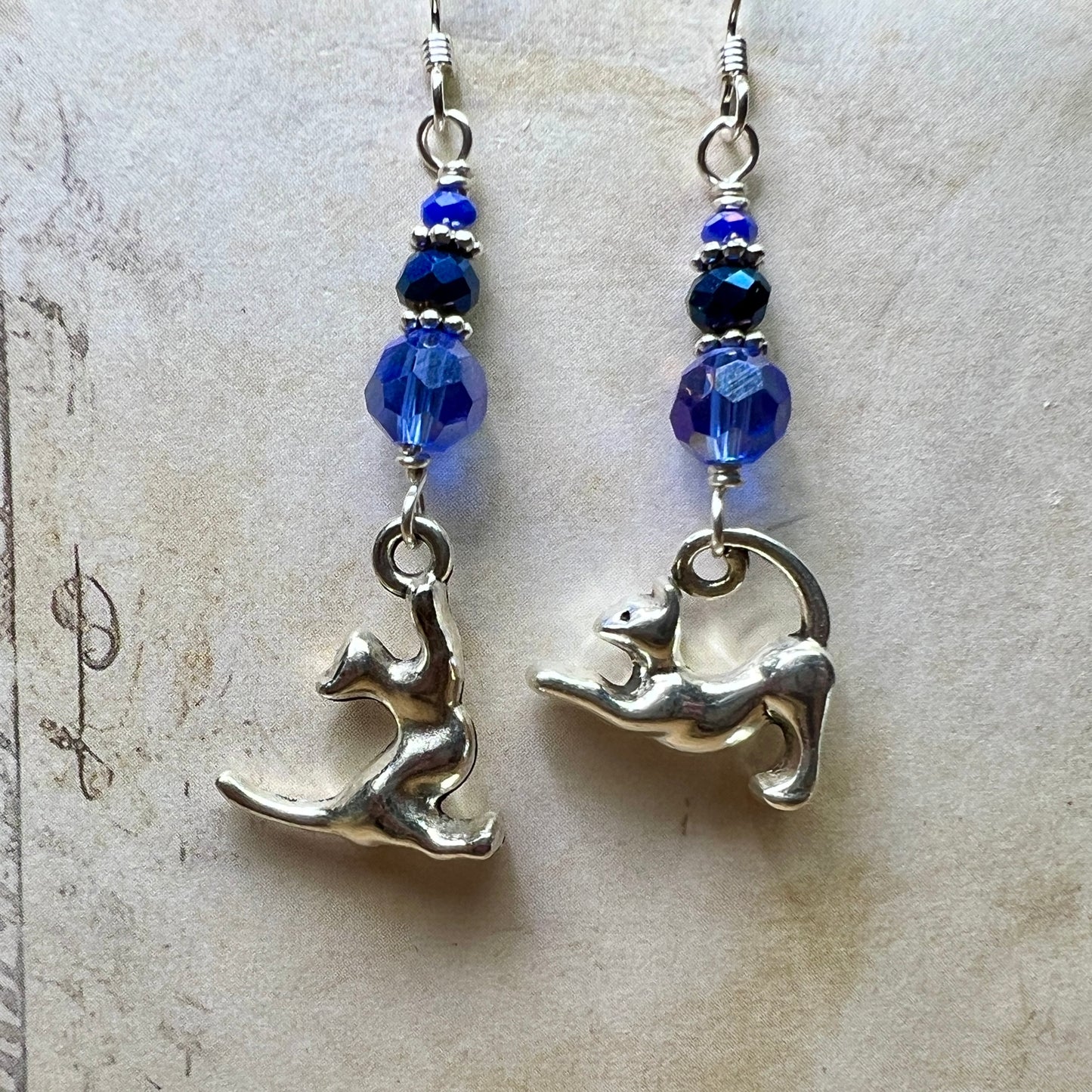 A pair of asymmetric earrings made from silvery kitty charms and three faceted glass beads in various shades of dark blue. The beads are graduated in size with thin silver spacers between them and the kitty charms swing freely below the beads. One kitty on its hind legs reaching  towards the beads with its front paws and the other is doing BIG STRETCH. 

In this photo they are hanging in front of an off-white paper backdrop. 