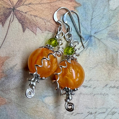 A pair of earrings that look like pumpkins, made from shimmery orange beads topped with smaller green glass beads with a tendril of silver wire spiraling down to the bottom of the earrings. 