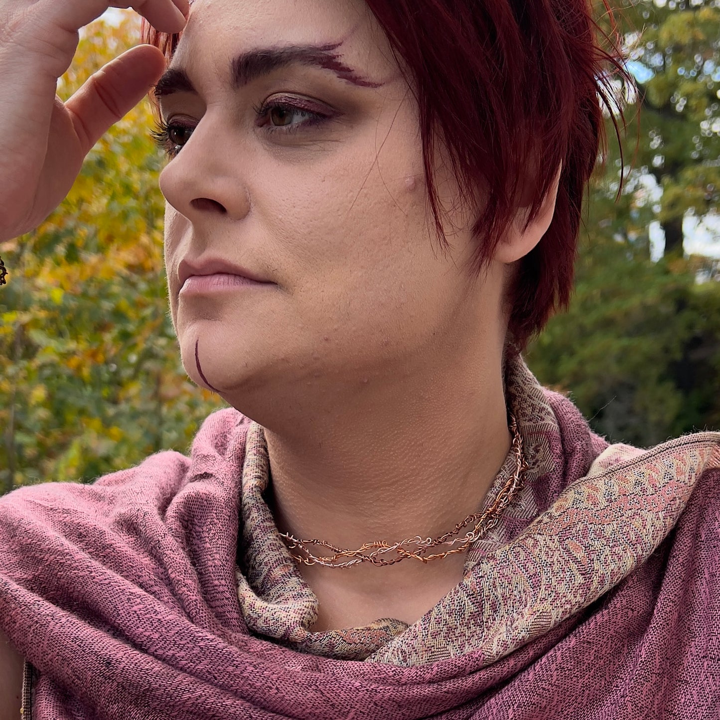 A light-skinned person with short red hair is wearing a mauve shawl and three neckvines braided together. They are pushing their hair out of their eyes.