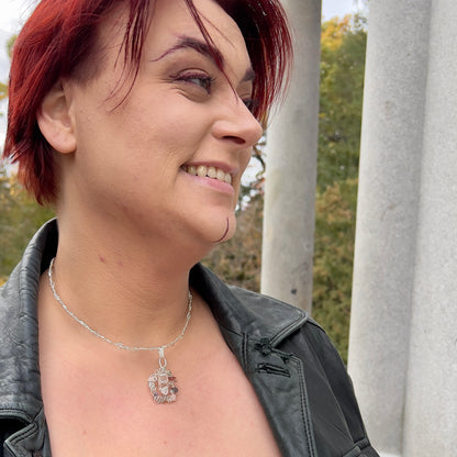 A light-skinned person with short red hair is looking away from the camera and smiling, wearing a black leather jacket and a silver neckvine with an intent pendant. 
