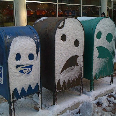 Three snow-covered USPS mailboxes standing outdoors in the snow in front of a building with large windows.